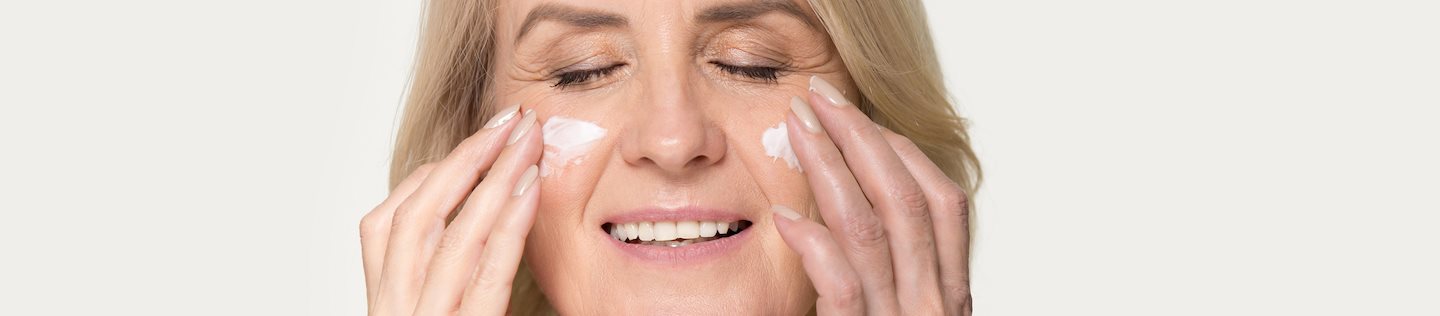 Une femme mûre souriante, les yeux fermés, masse délicatement un produit de soin sur ses joues du bout des doigts, illustrant ainsi une routine de soin anti-âge apaisante.