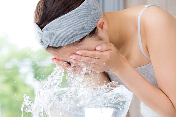 Frau mit grauem Haarband wäscht ihr Gesicht mit spritzendem Wasser.