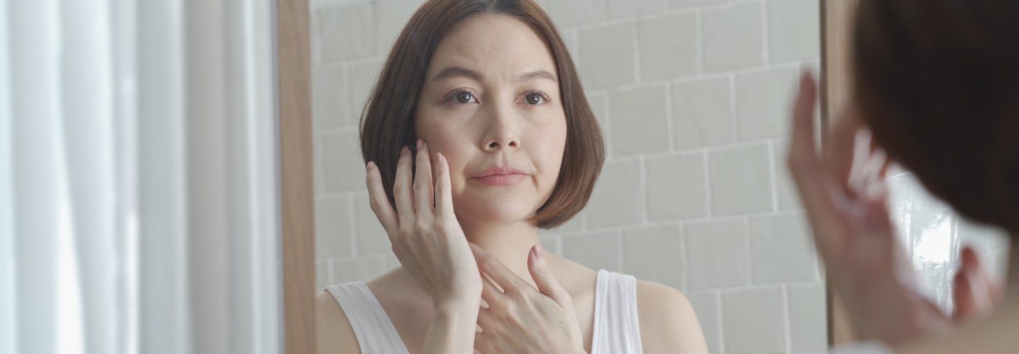 Asiatische Frau mittleren Alters betrachtet ihr Gesicht im Spiegel
