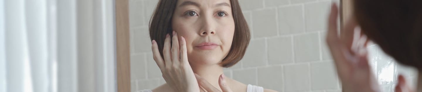 Asiatische Frau mittleren Alters betrachtet ihr Gesicht im Spiegel