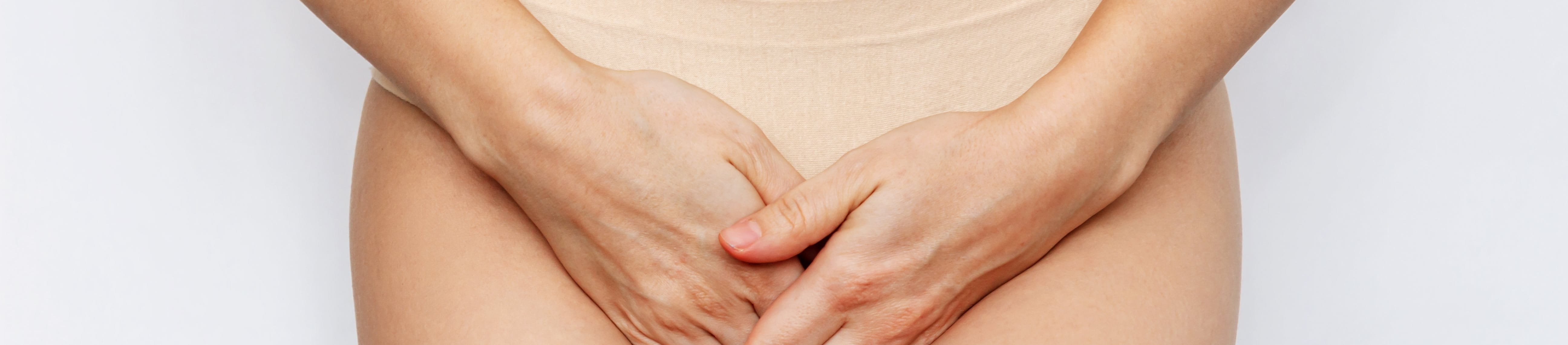 Frau hält sich ihre Hände schützend vor ihren Intimbereich. Sie trägt eine hautfarbene Unterhose und steht vor einem weißen Hintergrund