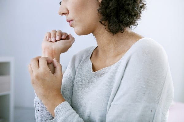 Eine Frau, in heller Kleidung, vor hellem Hintergrund, kratzt sich am Handgelenk.