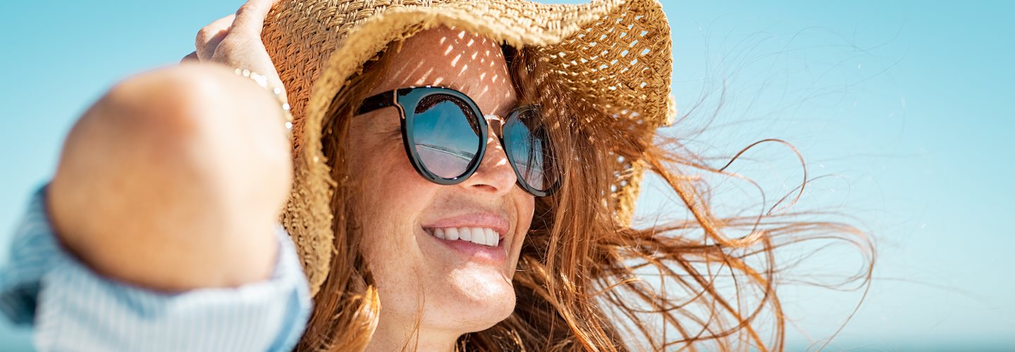 Rothaarige, lächelnde Frau mit Sonnenbrille und Sonnenhut am Strand