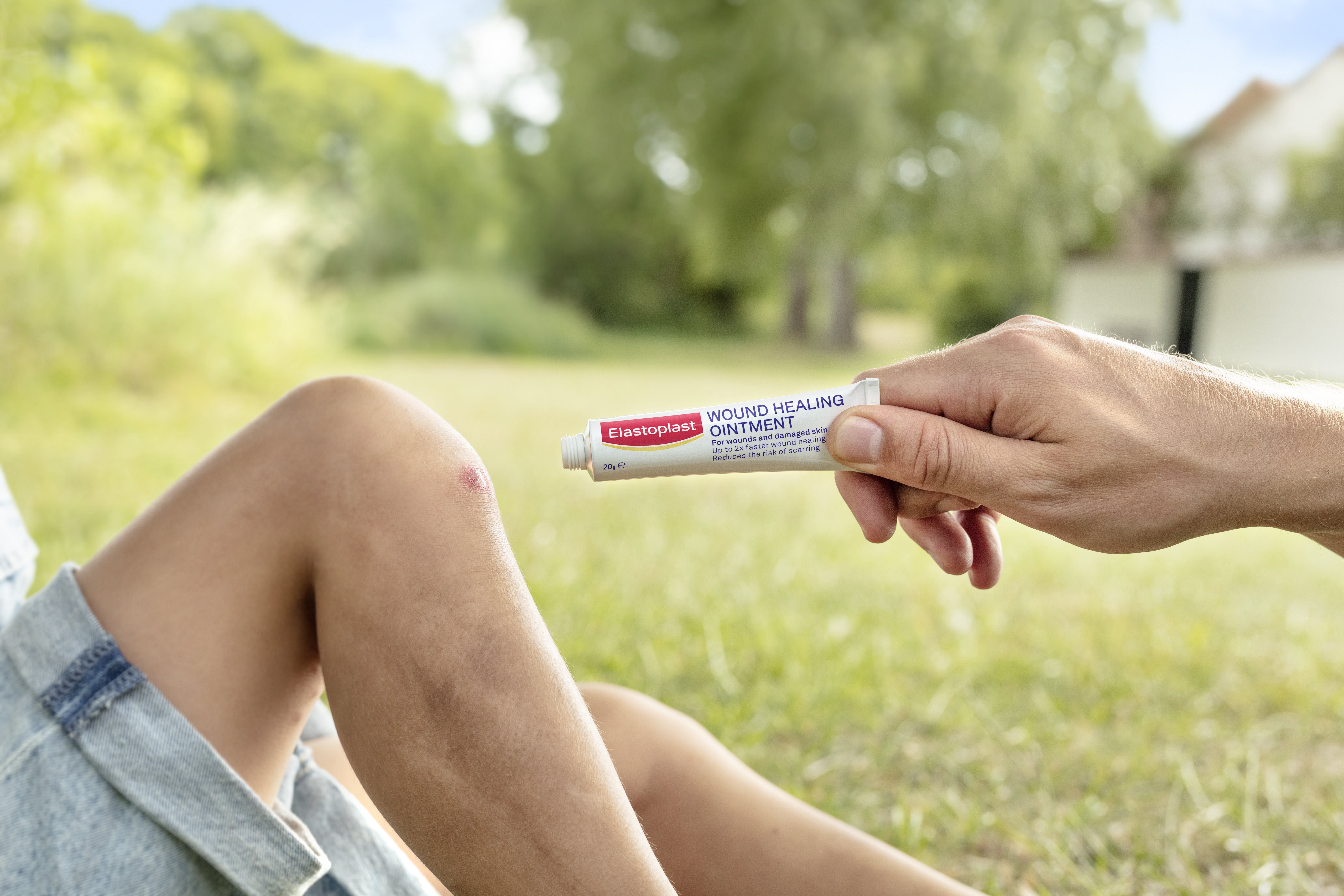 A parent's hand carefully applies Elastoplast Wound Healing Ointment to a child's wounded knee, promoting healing as the third and final step in pediatric wound care.