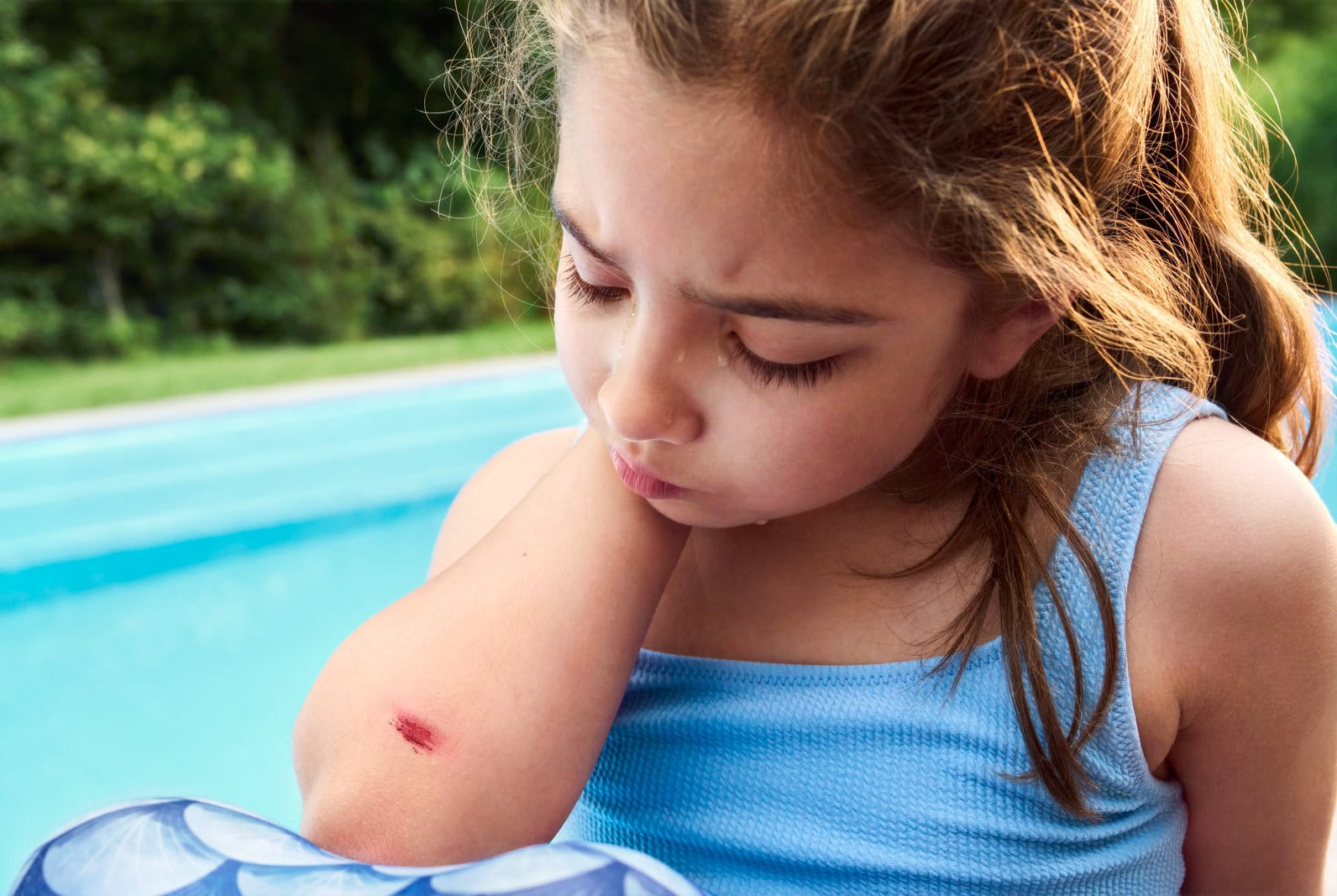 A young girl in a blue top worriedly examining a small, bloodied cut and scratch on her elbow, possibly from playing outside.