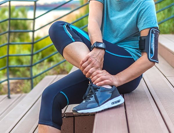 Person in Sportklamotten, die auf einer Treppenstufe sitzt und sich fest an ihr Fussgelenk greift.