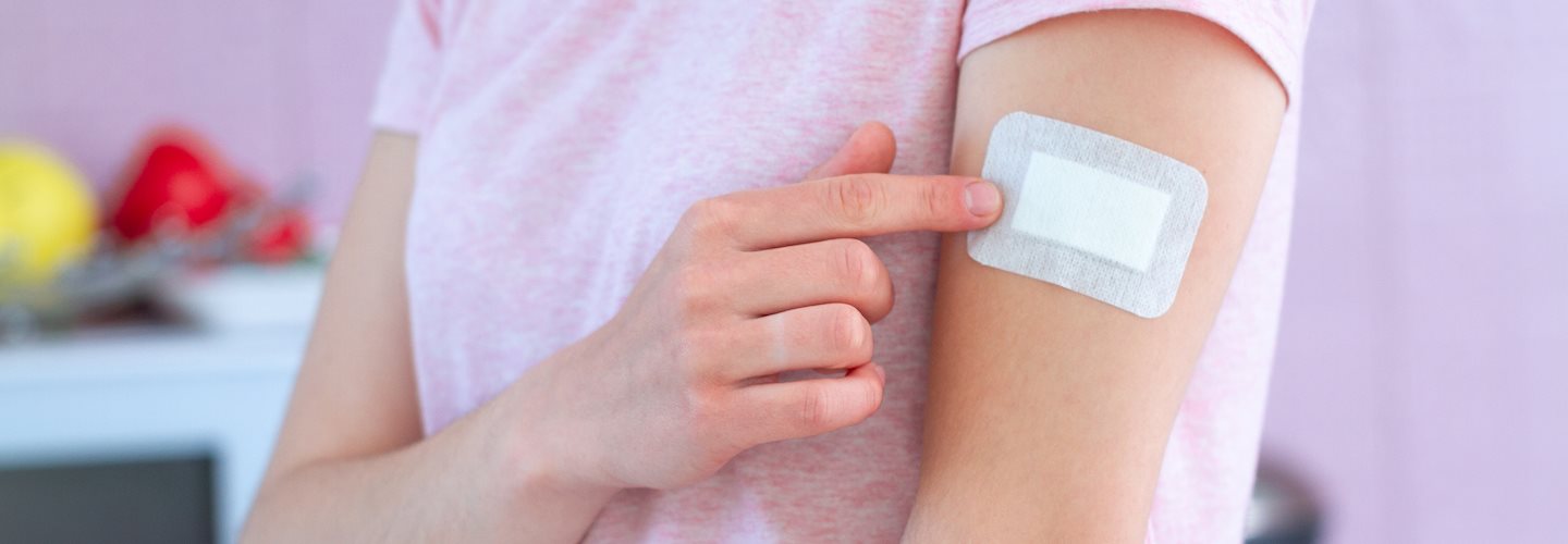 Eine Frau im pinken T-Shirt zeigt auf ihr Pflaster am Arm