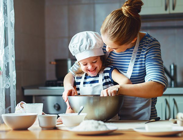 EIne Frau steht hinter einem Kind mit Kochmütze, während sie ihr dabei hilft, etwas in einer Schüssel umzurühren.