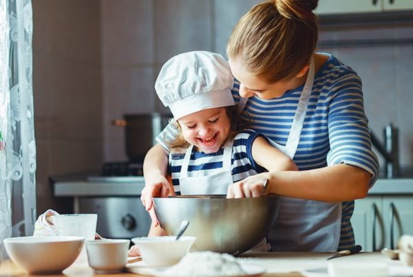 EIne Frau steht hinter einem Kind mit Kochmütze, während sie ihr dabei hilft, etwas in einer Schüssel umzurühren.