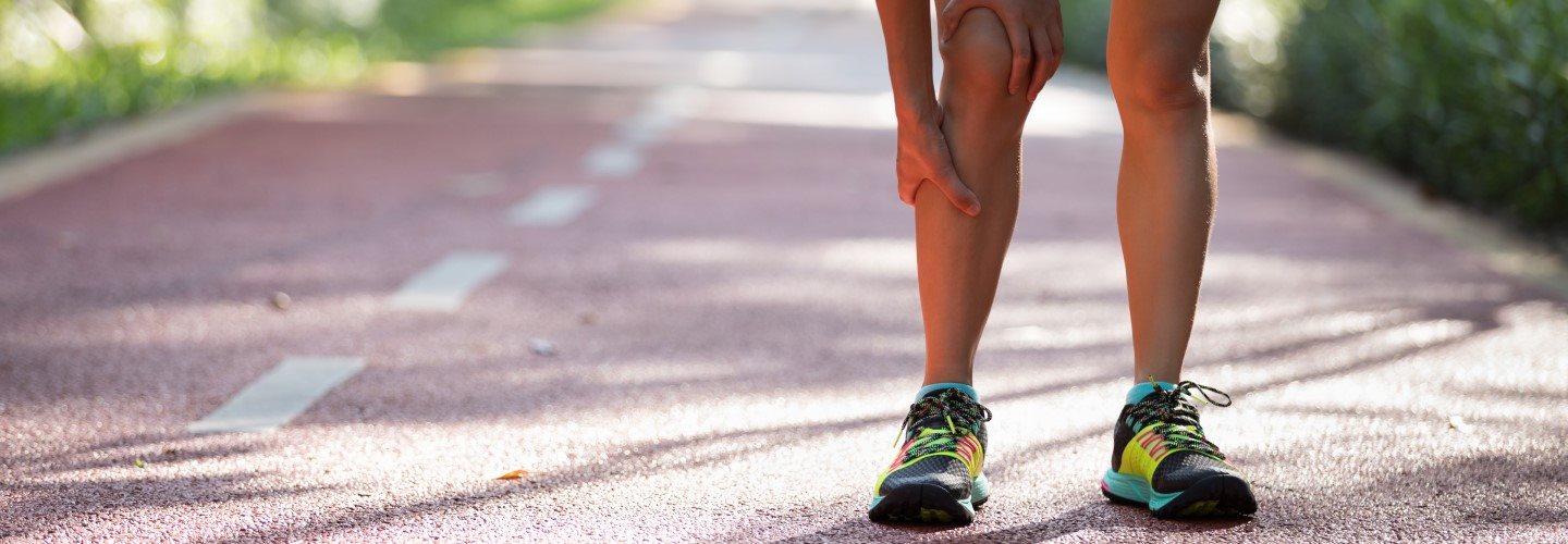Eine Frau im Sportoutfit steht vornüber gebeugt auf einer Landstraße und hält sich ihr schmerzendes Läuferknie. 