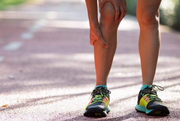 Eine Frau im Sportoutfit steht vornüber gebeugt auf einer Landstraße und hält sich ihr schmerzendes Läuferknie.