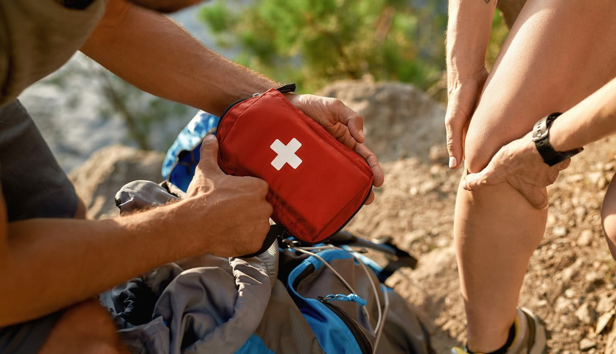 Mann hält Erste-Hilfe-Tasche in seinen Händen, während Frau sich ihr Knie vermutlich aufgrund von Schmerzen festhält. Sie befinden sich auf einem steinigen Gelände.