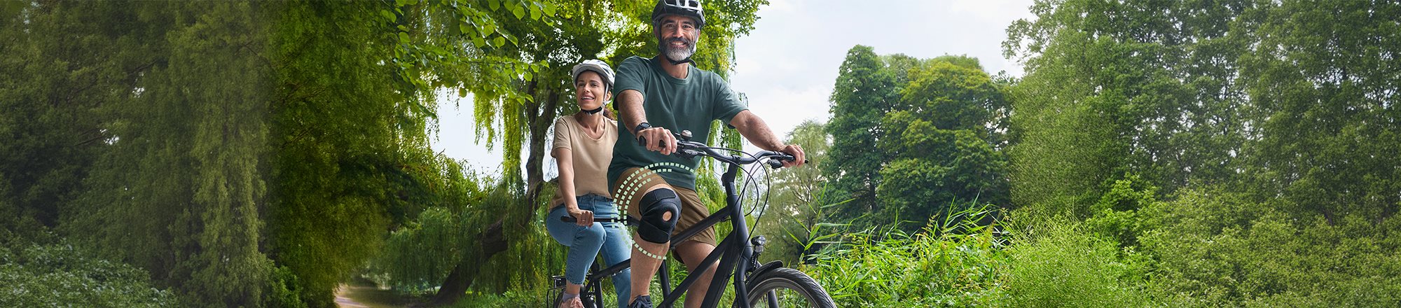 Two adults wearing bicycle helmets ride along a tree-lined path in a lush green park